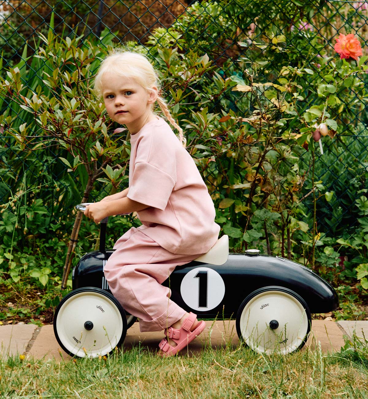 Child in pink T-shirt and trousers outfit riding a toy car outdoors with greenery in the background