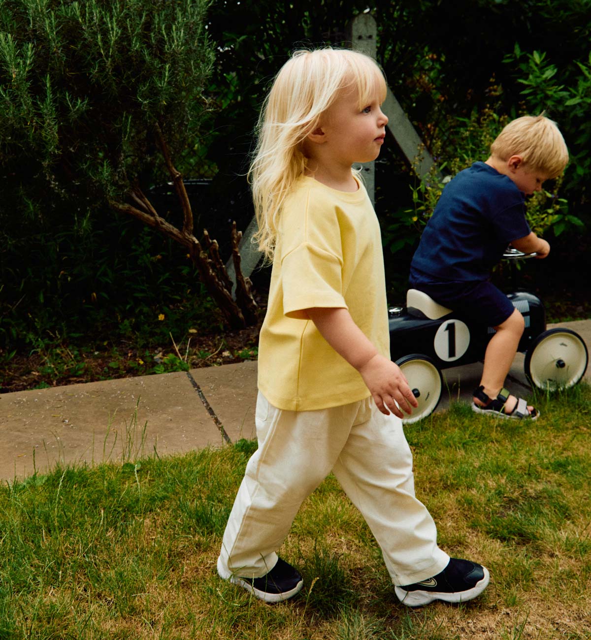 Two children playing outdoors, one in a yellow T-shirt and white trousers walking on grass, the other on a toy car wearing a navy T-shirt and navy shorts.